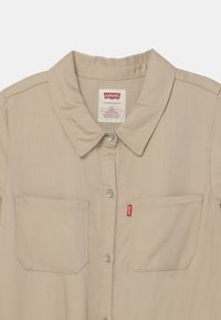 Beżowa koszula zapinana na guziki z kołnierzykiem, dwoma przednimi kieszeniami i metką Levi's. Posiada metalowe guziki oraz gładki, teksturowany materiał.