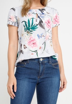 Kobieta w białej bluzce z krótkim rękawem w różowo-zielony kwiatowy wzór, połączonej z niebieskimi dżinsami.