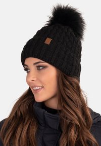 Bonnet en tricot noir avec un motif texturé, doté d'un pompon noir en peluche sur le dessus et d'une petite étiquette en cuir marron sur le côté.