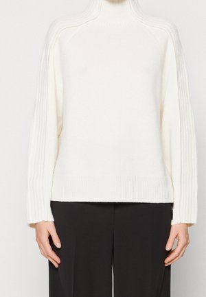 Weißer Strickpullover mit hohem Kragen, gerippten Ärmeln und Saum, der eine glatte Textur und eine lockere Passform aufweist. Kombiniert mit dunklen Hosen.