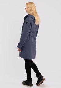Lång, marinblå vattentät jacka med huva, något kortad design. Bärs över svarta leggings och robusta svarta stövlar. Slät textur.