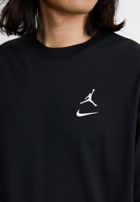 Melns kokvilnas T-krekls ar balto Jordana logotipu un Nike svītru kreisajā augšējā krūšu daļā. Īsas piedurknes un apaļas kakla izgriezums.