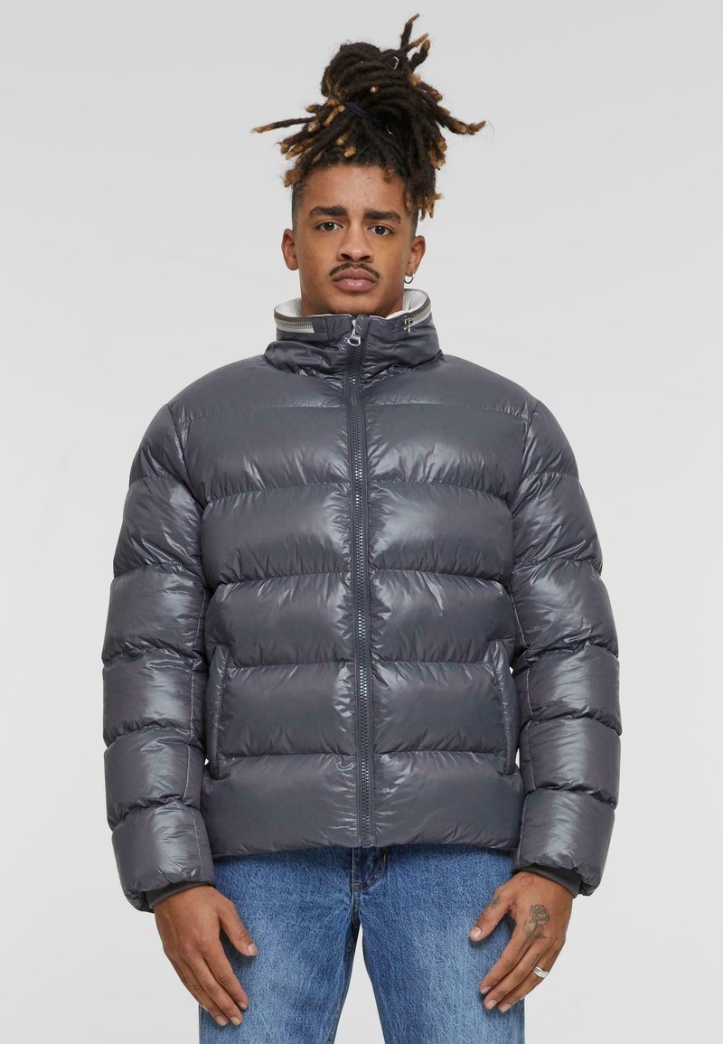 Grijze donsjack met gequilte secties, hoge kraag en ritssluiting. Het materiaal lijkt glanzend en geïsoleerd, gecombineerd met een broek in blauwe jeans.