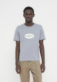 Ανοιχτό γκρι μπλουζάκι με στρογγυλή λαιμόκοψη και τυπωμένο οβάλ λογότυπο σε πράσινο και λευκό. Κοντά μανίκια, κατασκευασμένο από απαλό ύφασμα, συνδυασμένο με μπεζ παντελόνια.