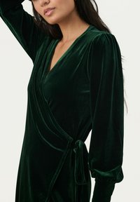 Grünes Samt-Wickelkleid mit langen Puffärmeln und tiefem V-Ausschnitt, ausgestattet mit einer gebundenen Taille und glatter Textur.