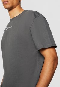 Tricou din bumbac gri, cu guler rotund, maneci scurte și un logo brodat alb subtil pe piept. Textură netedă, croială lejeră.