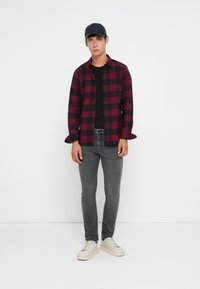 Joven de pie contra un fondo blanco, vistiendo una gorra negra, una camisa a cuadros roja y negra, una camiseta negra, jeans grises y zapatillas blancas.