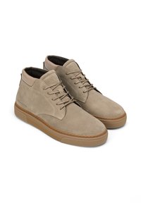 Baskets montantes en suede beige avec un système de laçage à l'avant, semelles en caoutchouc et cols matelassés, disposées côte à côte sur un fond blanc.
