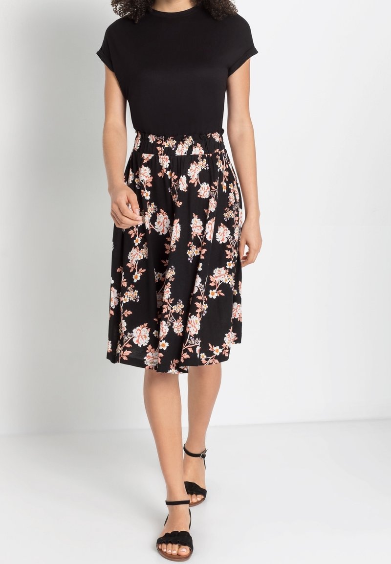 Femme portant un haut noir à manches courtes avec une jupe noire longueur genou à motif de fleurs roses et blanches, et des sandales noires à bride cheville.