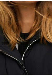 Schwarze Jacke mit einem Reißverschluss und silbernem Beschlag, kombiniert mit einem dunklen Sweatshirt. Der Stil betont Textur und Lagenlook.