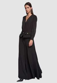 Tuta nera con maniche lunghe, scollo a V profondo, design plissettato nella parte anteriore, gambe ampie, vita stretta con cintura decorativa e bottoni dorati.