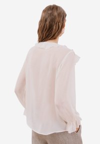 Bílá lehká halenka s volánovým límcem a dlouhými rukávy. Průsvitná textura a nařasené manžety. Spodní okraj mírně zakřivený vzadu.