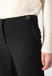 Pantaloni neri a sigaretta realizzati in un tessuto liscio, con tasche laterali e un logo in metallo decorativo in vita. Abbinati a un maglione crema.