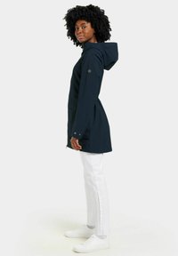 Marinblå vattentät parka med huva, sidofickor och knappdetaljer, i kombination med vita jeans och vita sneakers.