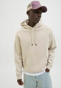 Beige Hoodie mit Kängurutasche, Kordelzug und gerippten Bündchen. Getragen mit einem weißen T-Shirt und hellblauen Jeans, ergänzt durch eine lila Mütze.