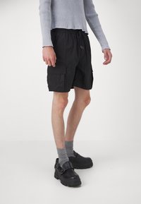 Shorts cargo noirs avec deux poches latérales, une taille élastique et un cordon de serrage. Portés avec un t-shirt à manches longues gris clair à côtes et des chaussettes grises.
