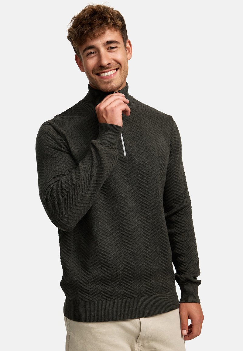Dunkelgrüner Pullover mit hohem Kragen, der ein strukturiertes Zickzackmuster, gerippte Bündchen und einen halben Reißverschluss aufweist.