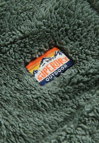 Fleece-Stoff in einem gedämpften Grünton, mit einem Patch, der orange und blaue Berge sowie den Text "SUPERDRY OUTDOOR" zeigt. Weiche, strukturierte Oberfläche.