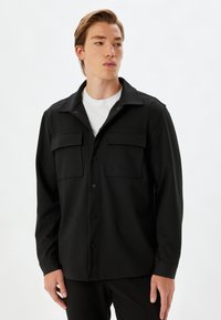 Camicia nera a maniche lunghe in tessuto testurizzato. Presenta due tasche sul petto, un colletto e una vestibilità rilassata. Maniche lunghe con polsini a bottoni.