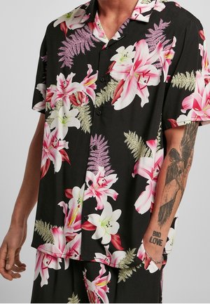 Man wearing black button-up shirt with pink and white lily flower and fern print, showing tattooed forearm with angel and text "BAD LOVE".