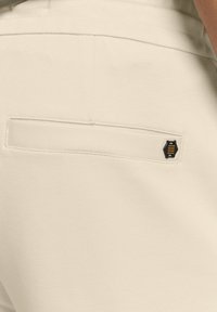 Beige broek met een gladde textuur, voorzien van een achterzak en een klein zwart en geel merklabel. Eenvoudig ontwerp, aansluitende pasvorm.