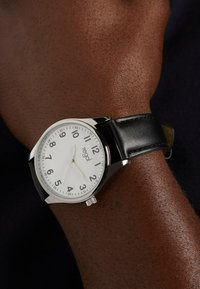 Round silver watch with a white dial, black leather strap, and large black numerals. Features a simple crown for time adjustment.