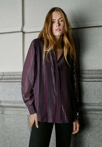 Blouse met lange mouwen in donkerpaars met verticale metalen strepen. Klassieke kraag, losse pasvorm en getextureerde stof. Gecombineerd met zwarte broek.