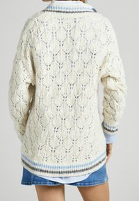 Cardigan crème tricoté avec un motif ajouré ouvert et des poignets côtelés. Présente des rayures horizontales bleues et grises à l'ourlet et au col.