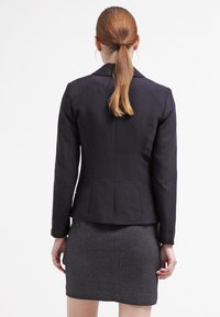 Granatowa dopasowana marynarka o wyraźnych ramionach, z pojedynczym wentylacją z tyłu i gładkim materiałem, zestawiona z teksturowaną szarą spódnicą.