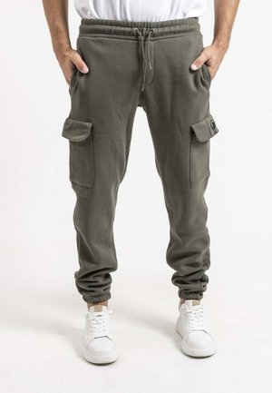 Homme portant un pantalon jogger cargo vert olive avec taille à cordon et des baskets blanches, mains dans les poches avant.