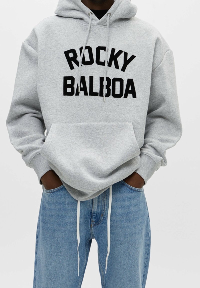 Personne portant un sweat à capuche gris clair avec l'inscription « ROCKY BALBOA » en lettres noires, les mains dans les poches, associée à un jean bleu clair avec des cordons blancs.