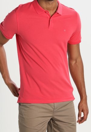 Hombre con una camisa polo roja brillante de manga corta y pantalones beige, de pie con una mano en la cadera.