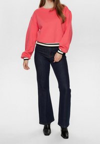Koralová mikina s oversized rukávy a černobílým pruhovaným lemem, spárována s tmavě modrými zvonovými džíny a černými kotníkovými botami.