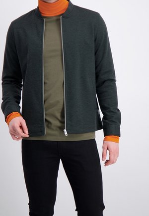 Mann trägt dunkelgrüne Zip-Up-Jacke über olivgrünem Hemd und orangefarbenem Rollkragenpullover, kombiniert mit schwarzen Hosen vor weißem Hintergrund.