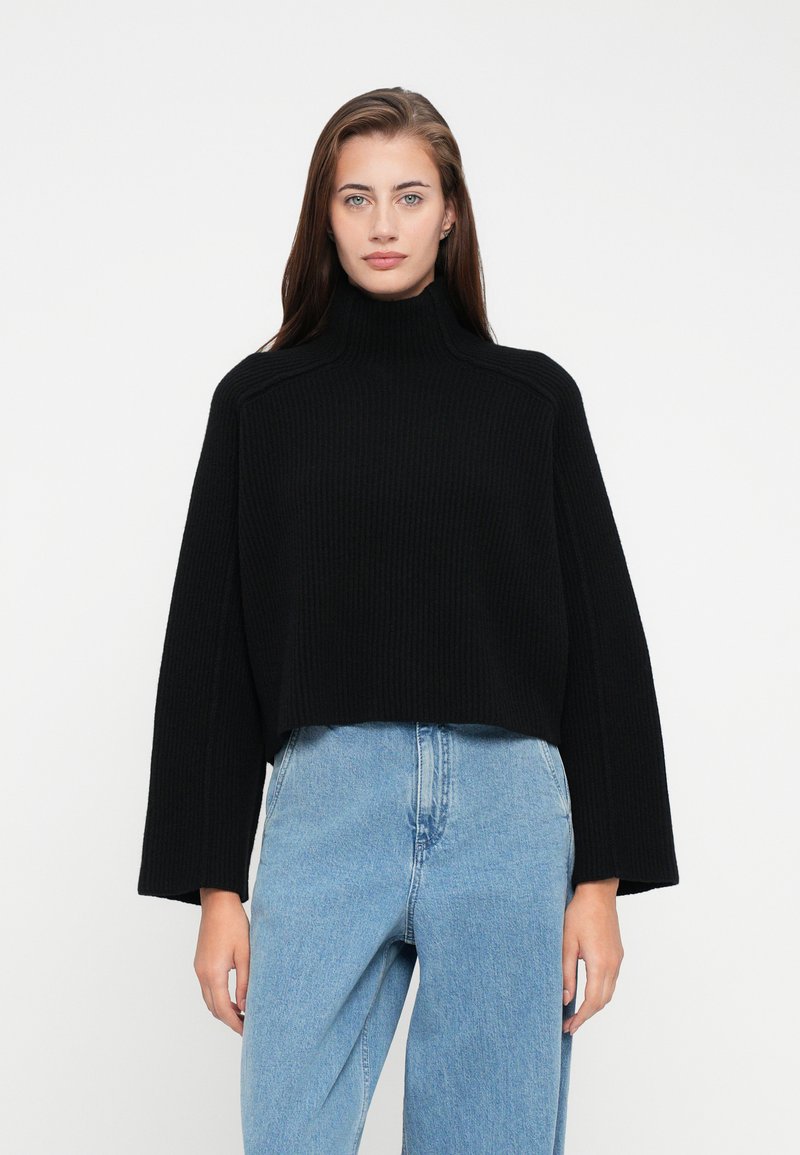 Woman with long brown hair wearing a black ribbed turtleneck sweater and light blue high-waisted jeans against a plain white background.