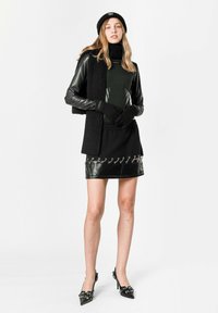 Chaqueta de cuero negro con bufanda de punto, vestido negro ajustado con detalles de imitación de cuero y herrajes plateados, y zapatos de tacón negros puntiagudos con detalles.
