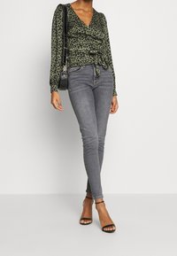 Top avvolgente con stampa leopardata verde, maniche lunghe, legato in vita, abbinato a jeans skinny grigi e tacchi neri con cinturini.