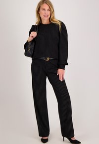 Zwart gestreept tweedelig pak met lange mouwen, elastische manchetten en broek met wijde pijpen. Draagt een riem en zwarte spitse pumps.