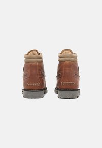 Bottes en cuir marron avec un col rembourré beige, dotées d'une semelle en caoutchouc texturée et de surpiqûres contrastantes à l'arrière.