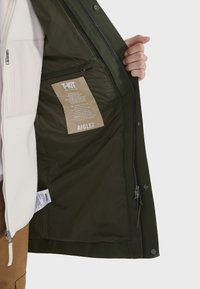 Grüne wasserdichte Jacke innen mit einem Etikett, einer Reißverschlusstasche und Fleece-Futter. Merkmale sind eine glatte Textur und sichtbare abnehmbare Teile.