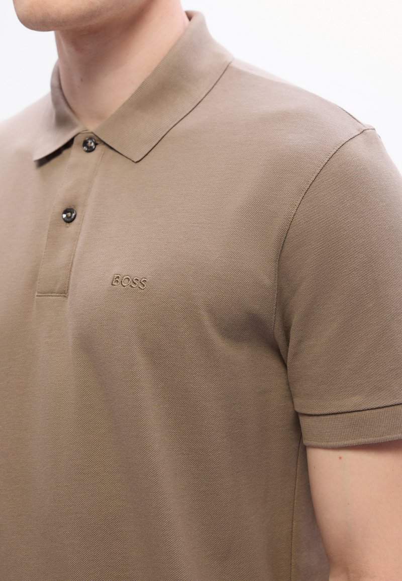 Polo shirt beige à manches courtes avec deux boutons noirs et logo "BOSS" brodé sur la poitrine gauche, porté par une personne.