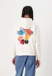 Cream hoodie with a rear graphic reading "OBEY STUDIOS" and colorful paint splotches in red, blue, yellow, and black. Soft fabric, casual fit.