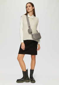 Grijze schoudertas met ronde vorm, gelaagd over een crème ribgebreide vest en witte blouse. Zwarte corduroy rok en chunky zwarte laarzen maken de look compleet.