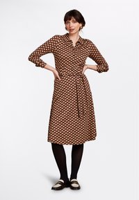 Abito camicia marrone con chiusura a bottoni e cintura in vita, caratterizzato da un motivo floreale rosso e crema. Maniche lunghe e lunghezza al ginocchio. Collant neri e mocassini.