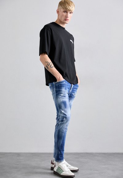 Zwarte oversized t-shirt met een ronde hals, blauwe distressed jeans met verfspetters en witte sneakers met groene strepen en kastanjebruine accenten.