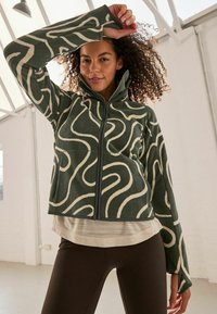 Groene fleecejas met witte abstracte patronen, hoge kraag en rits. Gedragen over een lichte top en zwarte leggings. Gestructureerde stof.