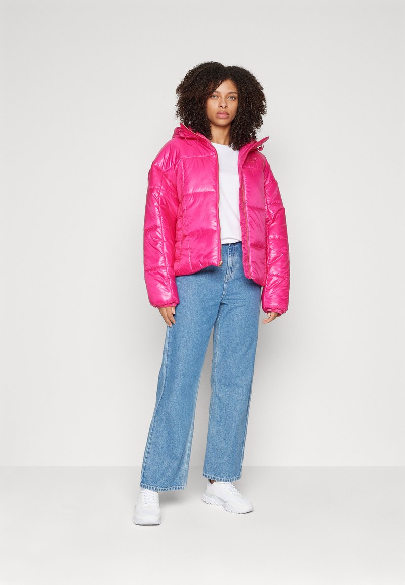 Rosa Pufferjacke mit glänzender Textur und Kapuze, kombiniert mit einem weißen T-Shirt und hellblauen Schlagjeans. Weiße Sneakers runden den Look ab.