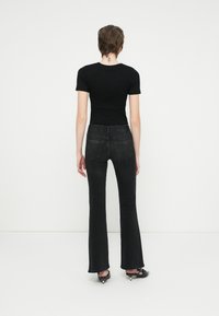 Camiseta negra de manga corta ajustada combinada con jeans negros de corte acampanado, que muestran bolsillos traseros y una textura suave. La persona lleva zapatos negros.
