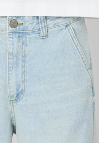 Jean en denim bleu clair avec une coupe large, dotés d'une fermeture à bouton, de poches avant et d'une texture légèrement délavée. Coutures jaunes accentuées visibles.