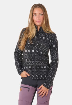Veste en polaire noire avec un motif de flocons de neige blancs, fermeture éclair intégrale, col haut et manches longues. Associée à un pantalon violet avec des zips.